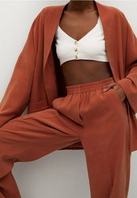 Rdzawokolorowy długi płaszcz, biały krótki top z guzikami oraz dopasowane spodnie z wysokim stanem. Gładka tekstura, luźny krój, minimalne detale w designie.