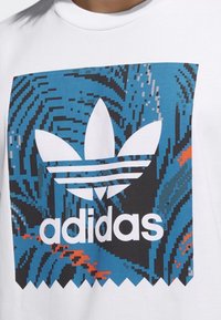 Vit t-shirt med ett blått och svart tryck med digitala mönster samt en överdimensionerad vit Adidas trefoil-logotyp, placerad centralt.