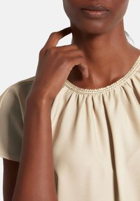 Beige Bluse mit gerafft gestaltetem Ausschnitt, strukturierten Details am Kragen und kurzen Ärmeln. Weiches Material mit glatter Oberfläche.