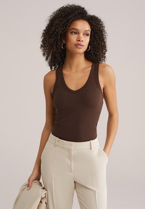 Göndör hajú nő ujjatlan barna felsőt és bézs nadrágot visel, egyik kezében bézs zakót tart, oldalra néz.