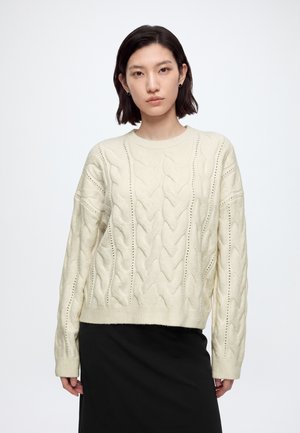 Femme avec des cheveux courts et foncés portant un pull torsadé crème et une jupe noire, debout devant un fond clair et uni.