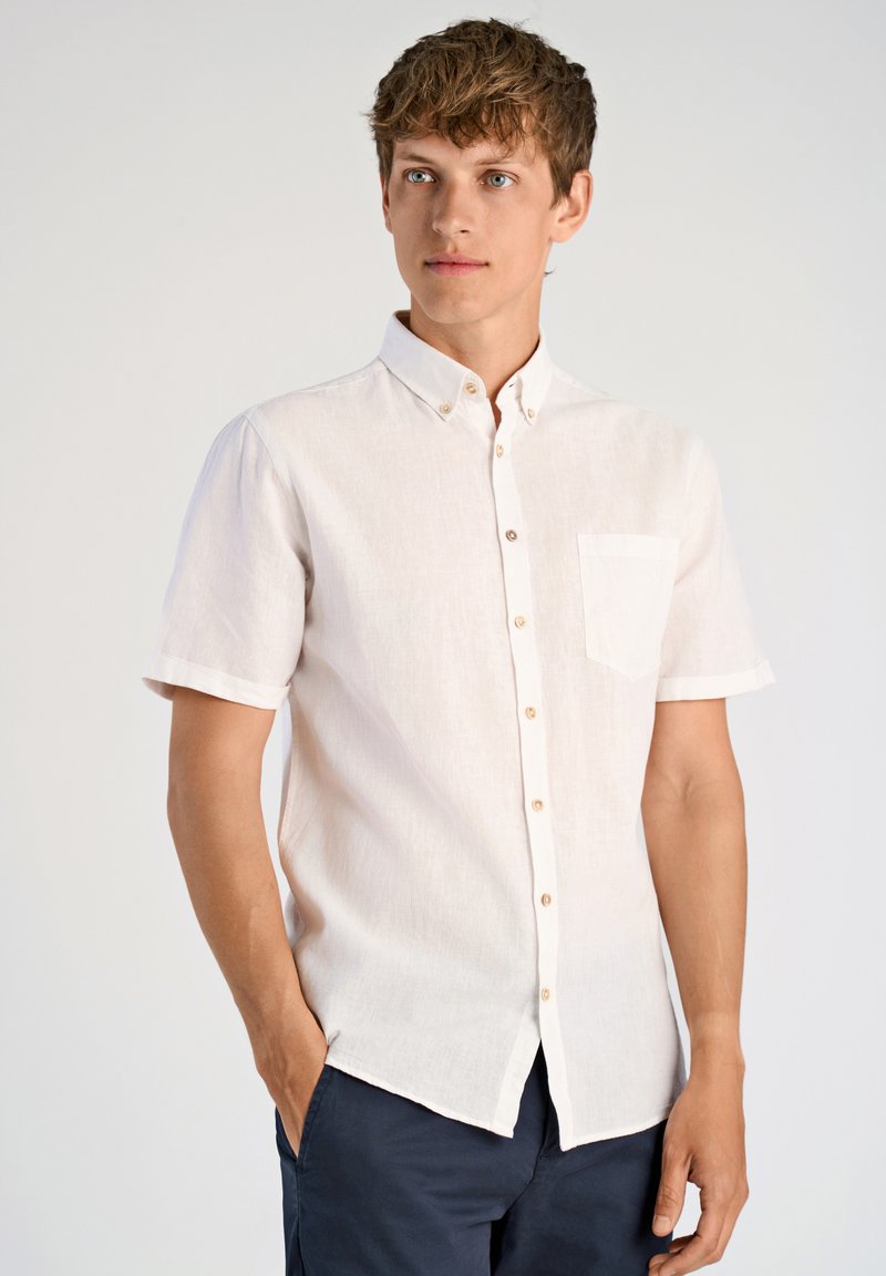 Camisa de lino de manga corta en color beige claro, con cuello abotonado, un bolsillo en el pecho y dobladillo redondeado. Los botones son de color marrón claro.