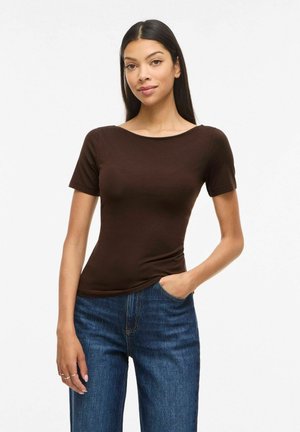 Femme aux longs cheveux foncés portant un haut marron à manches courtes ajusté et un jean bleu, une main dans la poche, debout devant un fond blanc.