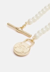 Pendentif en forme de cadenas doré avec des détails gravés, attaché à un fil de perles blanches brillantes. Présente un fermoir à bascule et des accessoires polis.