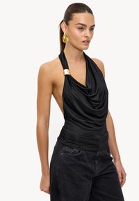 Haut noir à col licou avec un design drapé et des détails en métal doré, associé à un jean foncé. Le tissu semble lisse et ajusté.