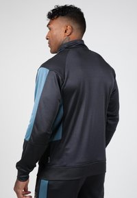 Zwart sportjack met blauwe accenten, hoge kraag en lange mouwen. Glad textuur met contrasterende zijpanelen.