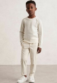 Kremast svetrom z rebrastim vzorcem in dolgimi rokavi, v kombinaciji s kremnimi hlačami po meri in belimi supergami. Gladka tekstura, prilagojen dizajn in minimalistični slog.