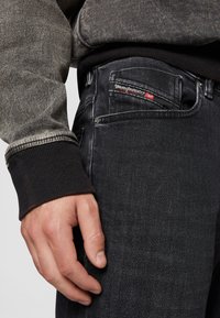 Zwarte spijkerbroeken van denim met een gestructureerde afwerking, voorzien van een zichtbare rode logo-tags, aangevuld met een grijze trui met een donkere geribde manchet.