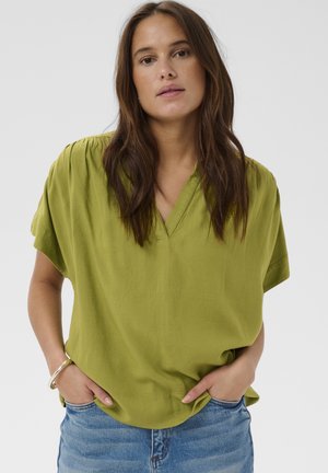 Femme aux longs cheveux bruns portant un chemisier vert clair à col en V et manches courtes, ainsi qu'un jean bleu, mains dans les poches, fond neutre.