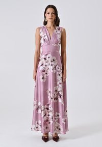 Frau trägt ein ärmelloses rosa blumiges Maxikleid mit tiefem V-Ausschnitt und burgundfarbene spitze Schuhe vor einem schlichten Hintergrund.