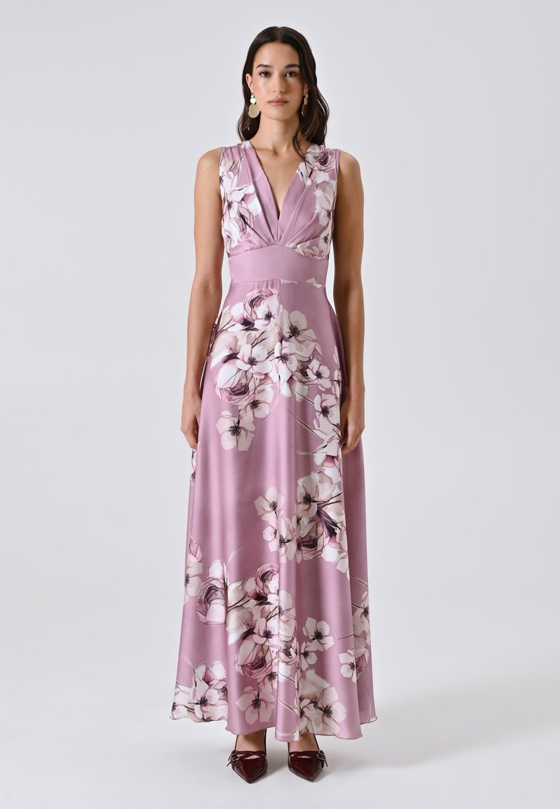 Frau trägt ein ärmelloses rosa blumiges Maxikleid mit tiefem V-Ausschnitt und burgundfarbene spitze Schuhe vor einem schlichten Hintergrund.