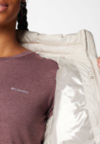 Giacca piumino leggera color beige con un'esterno liscio e dettagli in rete, indossata sopra una maglietta bordeaux con un piccolo logo.