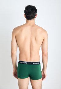Groene boxershorts met een zwarte tailleband waarop het witte "LACOSTE" logo staat, bekeken vanaf de achterkant. Glad stof en een aansluitend ontwerp.