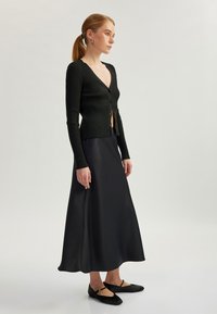 Schwarze gerippte Strickjacke mit tiefem V-Ausschnitt und Knöpfen, kombiniert mit einem fließenden schwarzen Satin-Maxirock und flachen Schuhen mit Knöchelriemen.