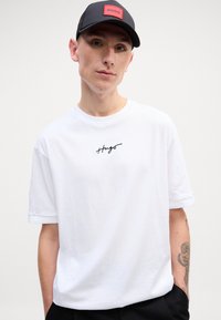 Hvid bomulds T-shirt med et sort Hugo-logo, rund halsudskæring og korte ærmer. Båret med en sort kasket med en rød logo-patch.
