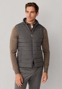 Grijze gewatteerde bodywarmer met een hoge kraag, een ritssluiting aan de voorkant en twee zijzakken, gedragen over een bruin shirt met lange mouwen, met subtiele stikpatronen.