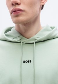 Φούτερ με κουκούλα σε ανοιχτό πράσινο χρώμα, κατασκευασμένο από μαλακό ύφασμα, με τσέπη καγκουρό και μαύρο λογότυπο "BOSS" στο στήθος, διαθέτει ρυθμιζόμενα κορδόνια.