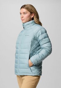Frau, die eine hellblaue, gesteppten Columbia-Jacke trägt, mit den Händen in den Taschen, zur Seite blickend, vor einem schlichten Hintergrund stehend.