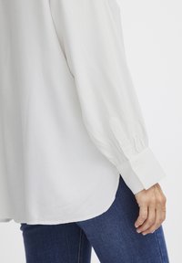Camicia bianca con tessuto testurizzato, maniche lunghe a sbuffo e polsini con bottoni, abbinata a jeans blu scuro. Orlo leggermente curvo nella parte inferiore.