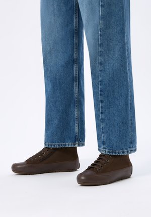 Persona che indossa jeans blu e scarpe casual marrone scuro con lacci, in piedi su sfondo bianco.