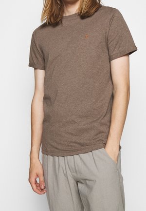 Osoba ubrana w brązowy krótki rękaw t-shirt z małym pomarańczowym haftem na piersi i jasnoszare luźne spodnie, ręka w kieszeni.