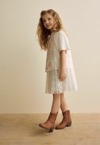 T-shirt graphique beige avec design floral, jupe blanche en dentelle superposée, et bottes chelsea marron à talon court et fermeture éclair latérale.