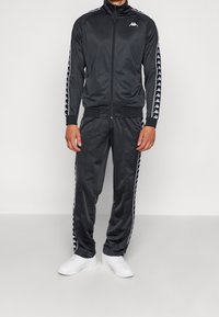 Homme portant un survêtement noir Kappa avec des bandes blanches du logo sur les manches et le pantalon, associé à des baskets blanches, debout devant un fond uni.