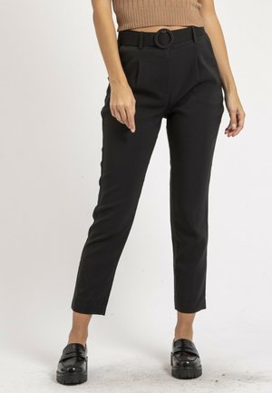 Femme portant un pantalon noir ajusté avec une ceinture à boucle ronde et des mocassins noirs, debout devant un fond uni.