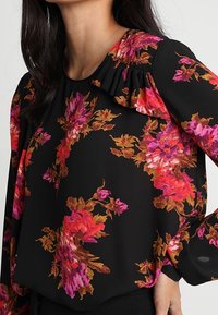Blusa nera con un motivo floreale in tonalità di rosa e arancione. Presenta un colletto arricciato e maniche lunghe. Tessuto leggero e liscio.