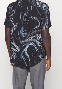 Uomo che indossa pantaloni grigi e una camicia nera a maniche corte con motivi astratti blu e bianchi, visto da dietro.