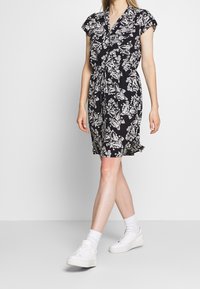 Robe noire à manches courtes avec un motif floral blanc, encolure en V et taille ajustable avec un lien. Portée avec des baskets blanches et des chaussettes.