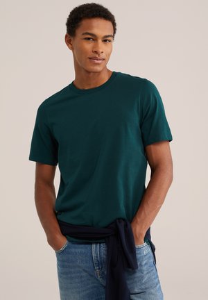 Jonge man die een donkergroen T-shirt met korte mouwen draagt, blauwe spijkerbroek en een marineblauwe trui om zijn middel gebonden, staand met zijn handen in de zakken.