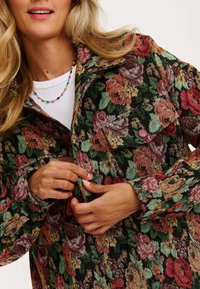 Kurtka w kwiatowy wzór w ciemnych odcieniach, z zielonymi, czerwonymi i różowymi różami. Posiada zapięcia na guziki i kieszenie na piersi, wykonana jest z teksturowanej tkaniny.