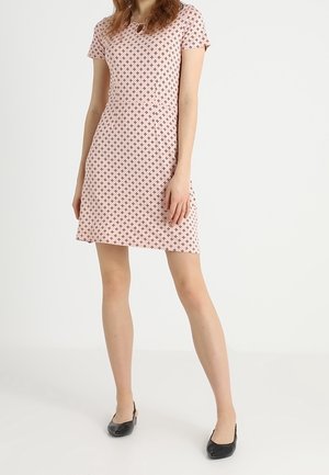 Frau in einem kurzärmeligen, knielangen rosa Kleid mit kleinem schwarzem Kreuzmuster, stehend mit schwarzen flachen Schuhen vor weißem Hintergrund.