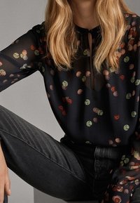 Kvinna som bär en svart genomskinlig blus med flerfärgade prickar och högmidjade mörkgrå jeans, sitter med ett ben böjt.