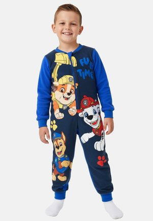 Smiling boy wearing blue Paw Patrol-themed onesie with characters Chase, Rubble, and Marshall, standing barefoot against a white background.