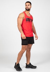 Rode mouwloze tanktop met zwart logo en tekst, gecombineerd met zwarte shorts en zwarte sportschoenen. Onderwerp staat in profiel.