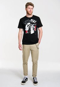 Black cotton t-shirt with "Spy vs Spy" graphic in white and red, paired with tan trousers; model wears black and white sneakers.