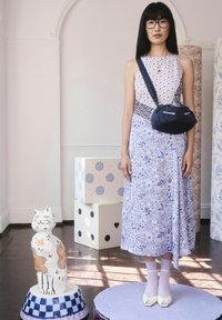 Modelka v šatách so vzorom s námorníckym crossbody kabelkou stojí vedľa keramickej sošky mačky. V pozadí sú dekoratívne krabice.