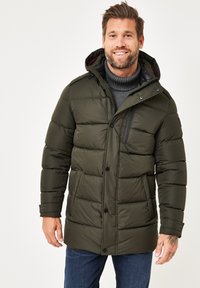 Dunkelgrüne Pufferjacke mit Kapuze, ausgestattet mit einer gesteppten Textur, schwarzen Reißverschlussdetails und Knöpfen. Getragen über einem grauen Rollkragenpulli.