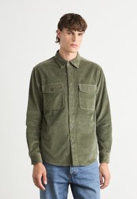 Olijfgroene corduroy shirt met een kraag, twee borstzakken en knoopsluitingen. Heeft een zachte textuur en een relaxte pasvorm.