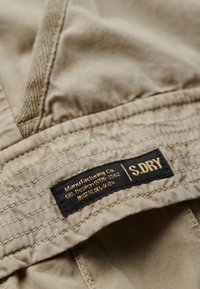Beige stof met een geborduurd patroon en zichtbare stiksels. Zwarte label met gouden tekst die "Manufacturing Co." en "S.DRY." accentueert.