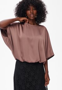 Vrouw met krullend haar, gekleed in een losse mauve blouse en een zwarte kanten rok, poseert met één hand bij haar hals tegen een witte achtergrond.