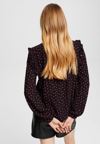 Schwarze Bluse mit rosa Punkten, mit Rüsche an den Schultern und langen Ärmeln. Das Material wirkt glatt; tailliert.