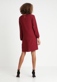 Robe bourgogne à manches longues, décolleté rond et détail en forme de trou de serrure au dos. Accents d'épaules ornés, tissu lisse et coupe légèrement évasée.