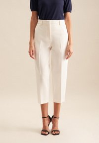 Weiße, maßgeschneiderte, cropped Hosen mit Falten, aus glattem Stoff gefertigt. Getragen mit einem marineblauen Oberteil und schwarzen Riemchen-Heels. Elegantes Design.