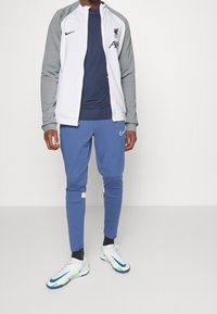 Chándal Nike que incluye una chaqueta de cremallera blanca y gris, camiseta interior azul marino, pantalones de chándal azules y zapatillas blancas con detalles en azul y amarillo.