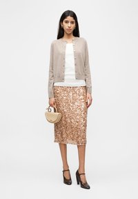 Vrouw draagt een beige cardigan, wit shirt, rosegouden midirok met pailletten, zwarte hakken en houdt een kleine beige handtas vast, terwijl ze tegen een witte achtergrond staat.
