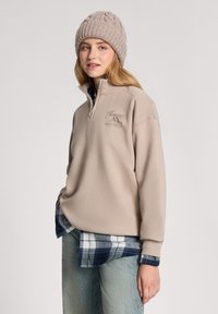 Beżowy polarowy sweter z półzamkiem, z haftowanymi grafikami górskimi. Nosi się go na niebiesko-białą koszulę w kratę, w połączeniu z jasnymi dżinsami.