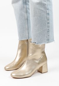 Bottines métalliques dorées avec une texture lisse, un bout pointu et un talon bloc. Portées avec un jean bleu clair usé qui s'arrête au-dessus de la cheville.
